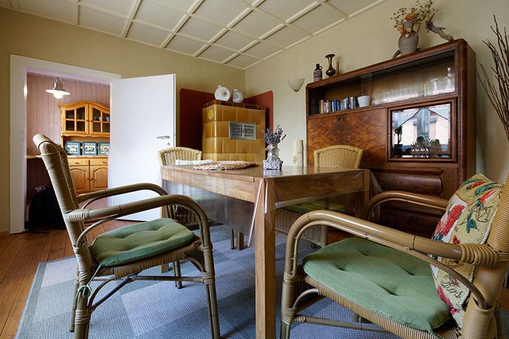 Holzesstisch mit vier Armlehnstühlen, dunkler Holzschrank, helle lehmverputzte Wände, hellbrauner Heißluftkachelofen, aufgearbeiteter Dielenfußboden.